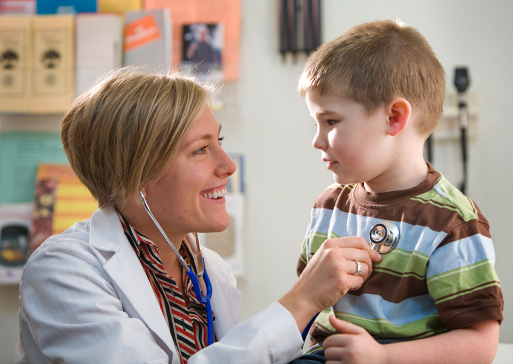Female doctor in white coat with stethoscope examining smiling boy in striped shirt.