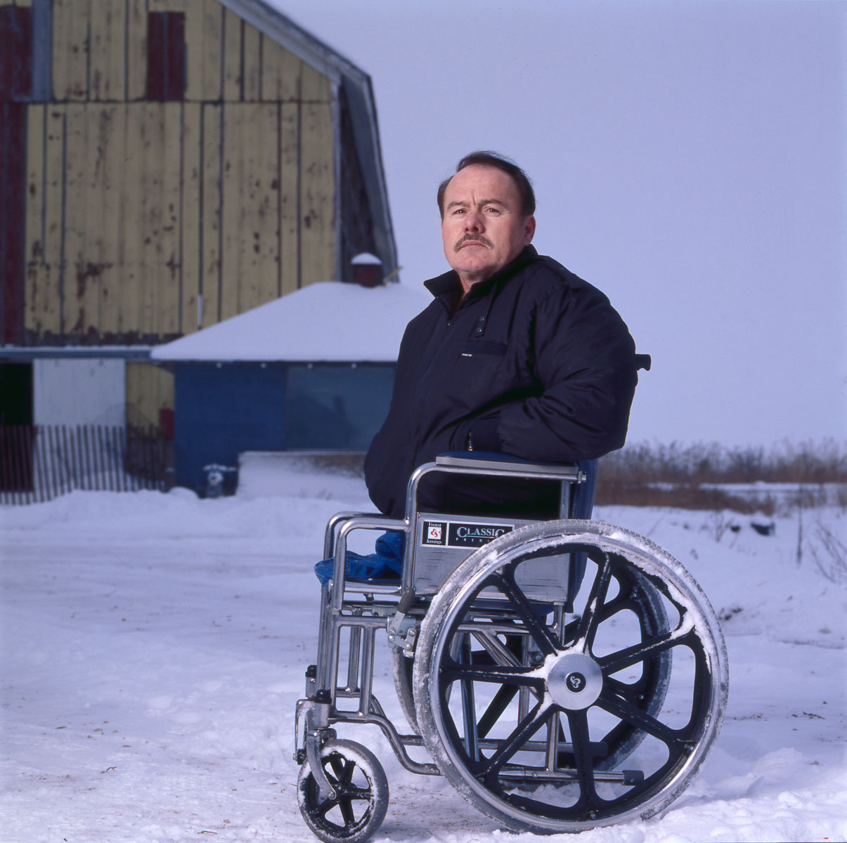Man in wheelchair sitting in front of snowy barn.