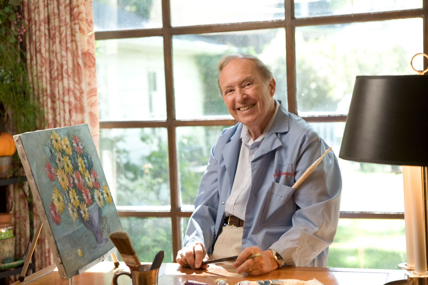 Artistic man in blue jacket painting at a desk in front of window.