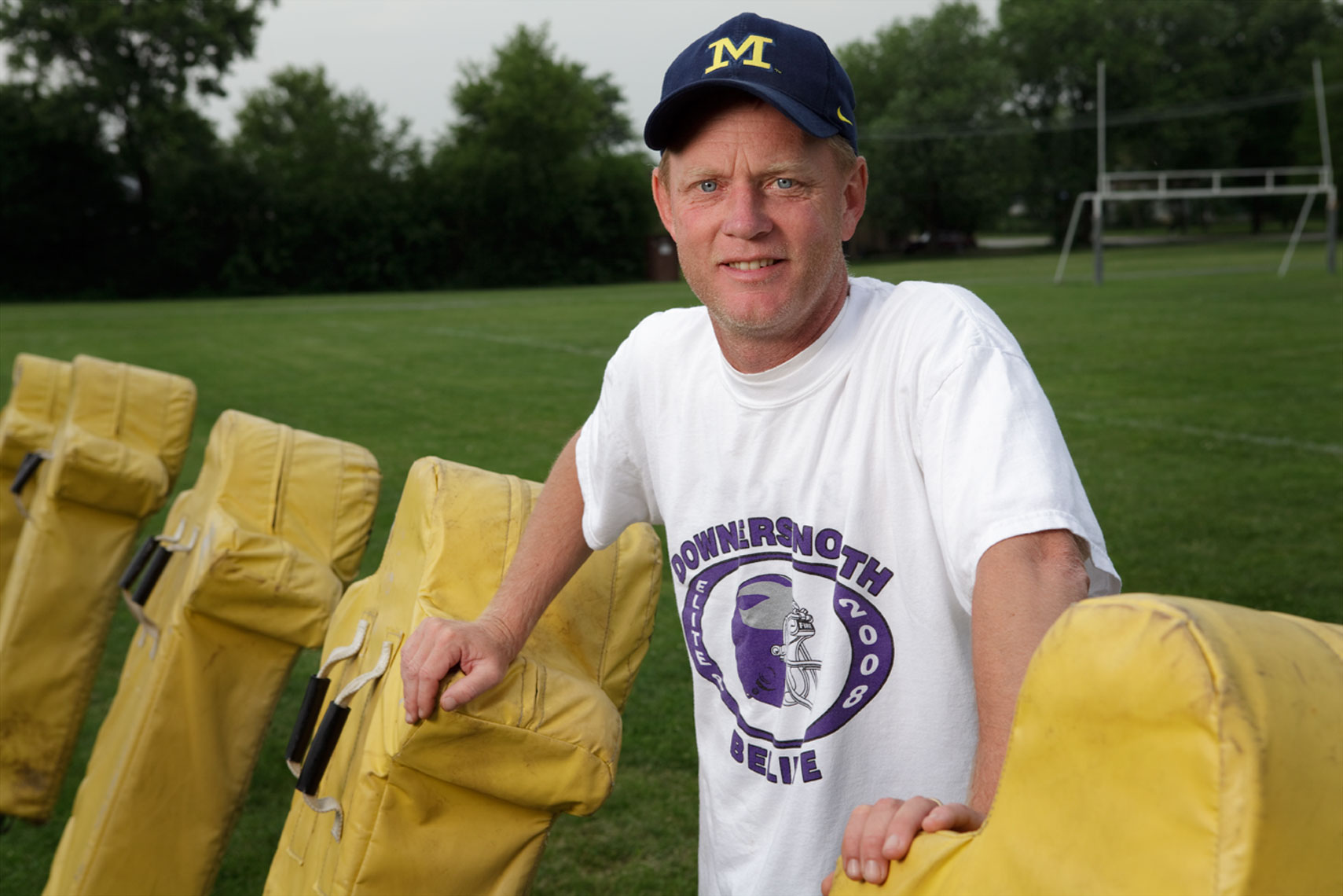 High school football coach with University of Michigan hat standing by yellow blocking sled.